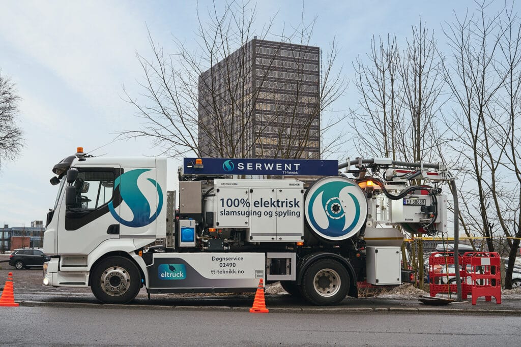 En stor hvit elektrisk lastebil med "100% elektrisk slamsuging og spyling" skrevet på siden står parkert på en gate. Oransje kjegler omgir den. En høy bygning er synlig i bakgrunnen, sammen med bladløse trær.