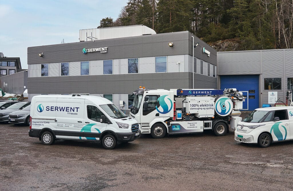 Hvite nyttekjøretøy med «SERWENT»-merke står parkert utenfor et moderne, grått industribygg med store vinduer. En blå garasjeport er synlig, sammen med flere trær i bakgrunnen.