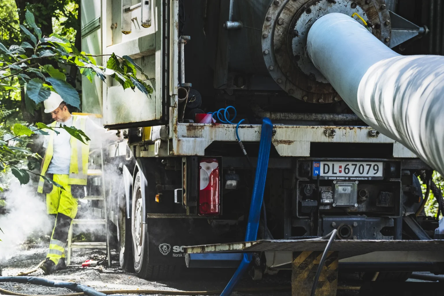 En arbeider i synlige klær og hjelm står ved en stor rørfornying bruksbil med rør, omgitt av damp eller røyk, på en solskinnsdag. Bilens skilt viser DL 67079.