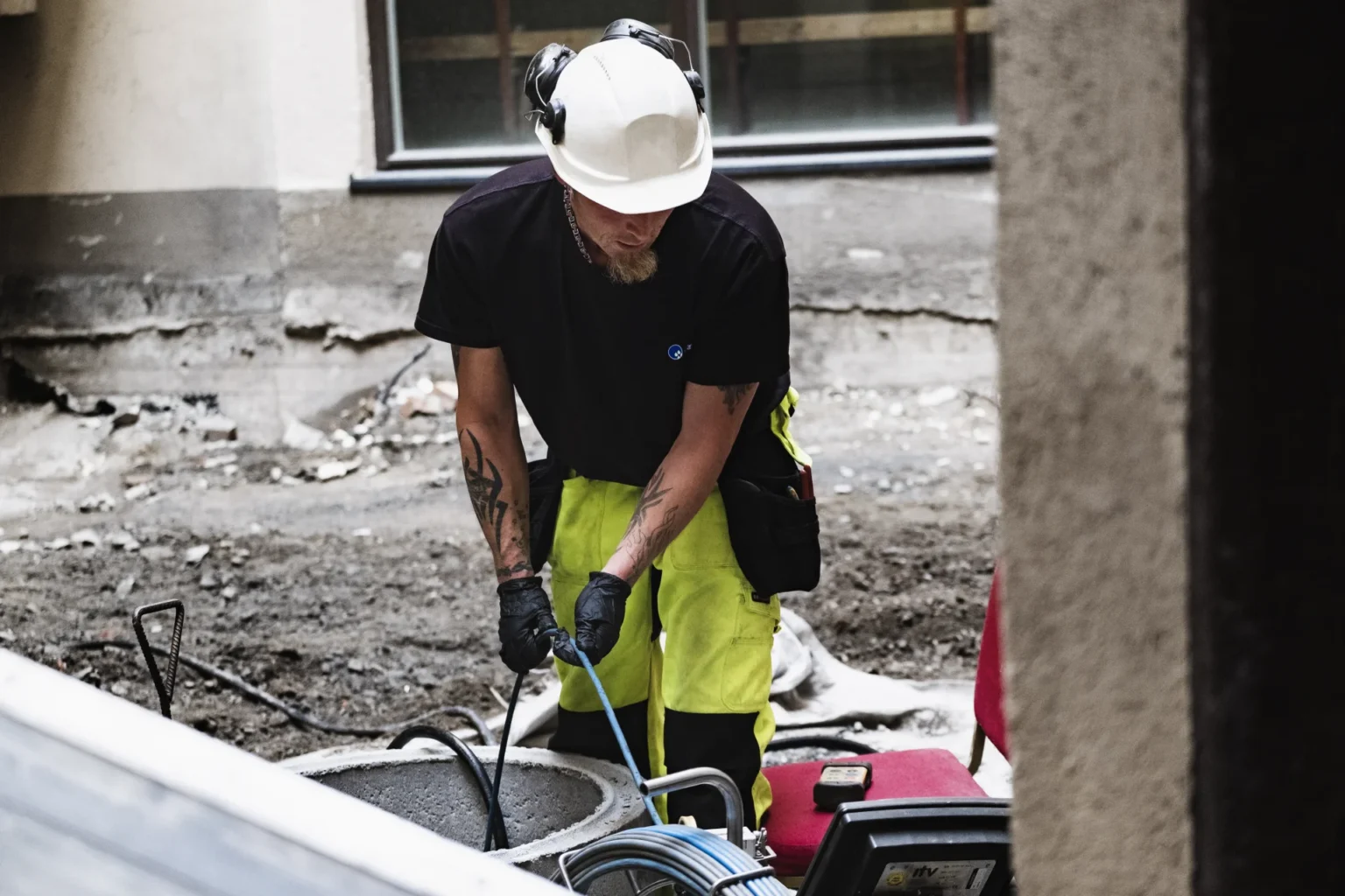 En bygningsarbeider iført hvit hjelme, hørselvern, svarte hansker og synlige gule bukser håndterer kabler for høytrykksspyling på en arbeidsplass med synlig jord og bygningsvegger i bakgrunnen.