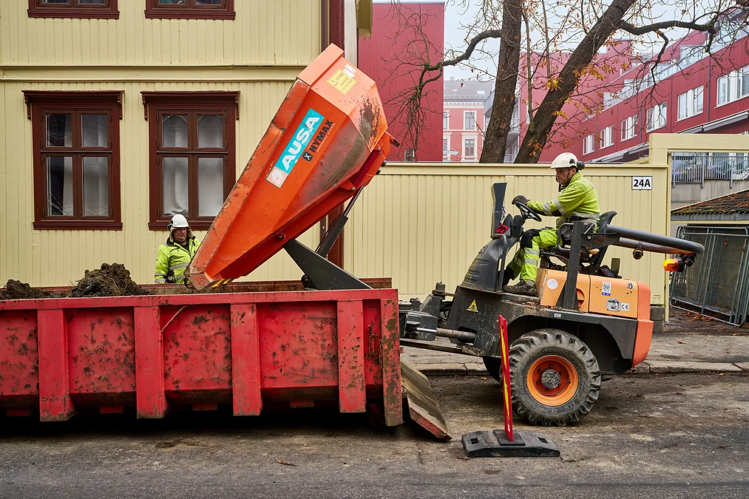 To Serwent-bygningsarbeidere i sikkerhetsutstyr betjener en oransje AUSA-dumper, og losser jord i en stor rød container ved siden av en gul bygning i en bygate.