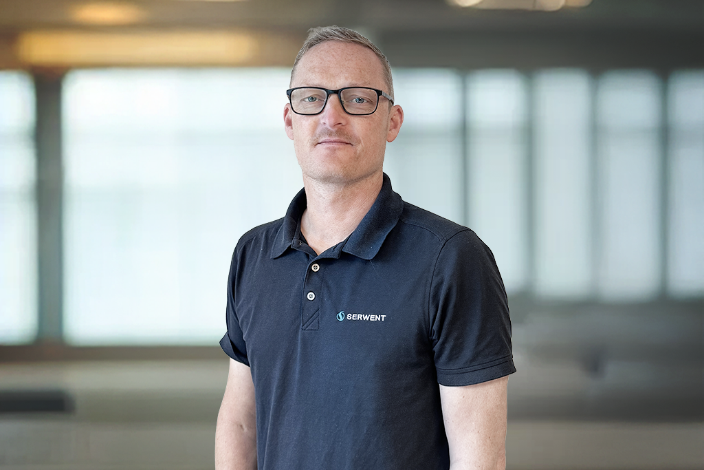 En mann med kort, blondt hår og briller, iført svart poloskjorte med SERWENT-logo, står innendørs foran store vinduer med uskarp bakgrunn.