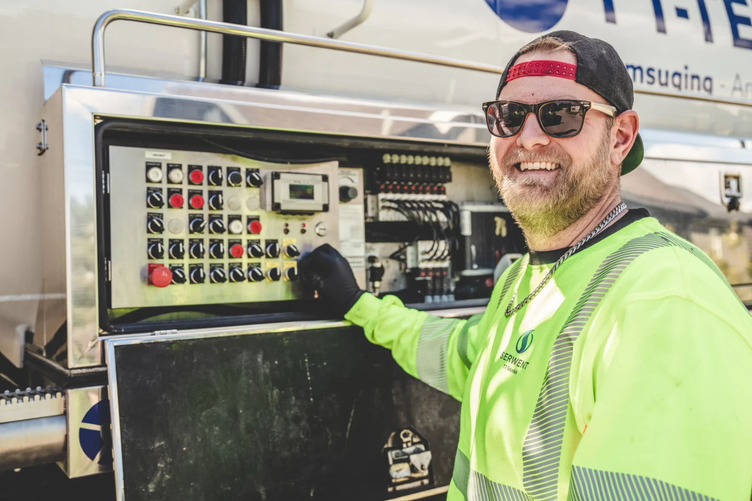 En mann i neon sikkerhetsskjorte, hansker og solbriller smiler mens han betjener et kontrollpanel på industriutstyr utendørs, og viser frem sin ekspertise med ADR-tjenester.