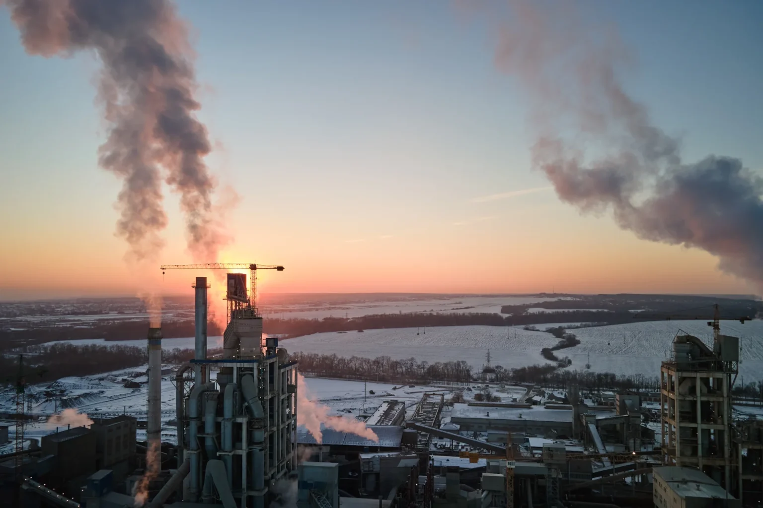 Industrianlegg med høye skorsteiner som slipper ut røyk, plassert mot et vinterlandskap med snødekte jorder og fjerne trær ved soloppgang eller solnedgang. Himmelen er klar, og sollyset filtrerer gjennom røyken.