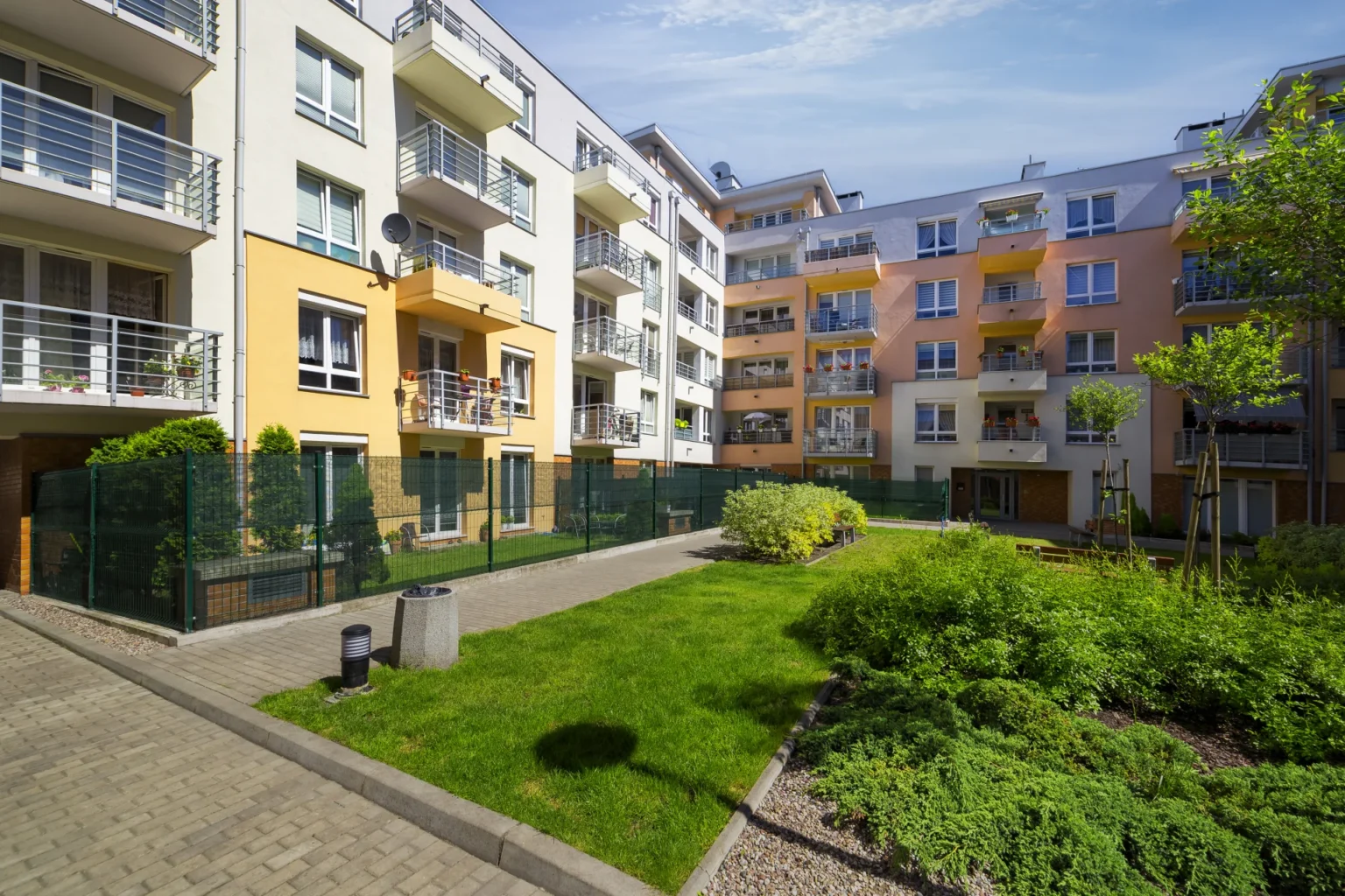 Moderne Serwent-leilighetsbygg med balkonger omgir en velholdt gårdsplass med grønt gress, busker og unge trær, under en delvis skyet blå himmel.
