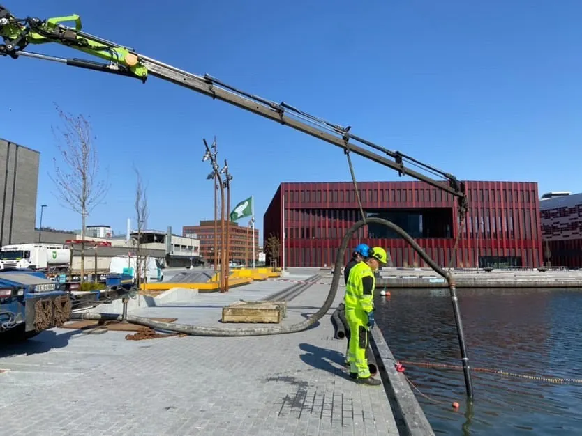 To arbeidere i lyse verneutstyr betjener en lang, kranlignende maskin med en slange som strekker seg inn i en vannmasse på en kai, med moderne bygninger og klarblå himmel i bakgrunnen.