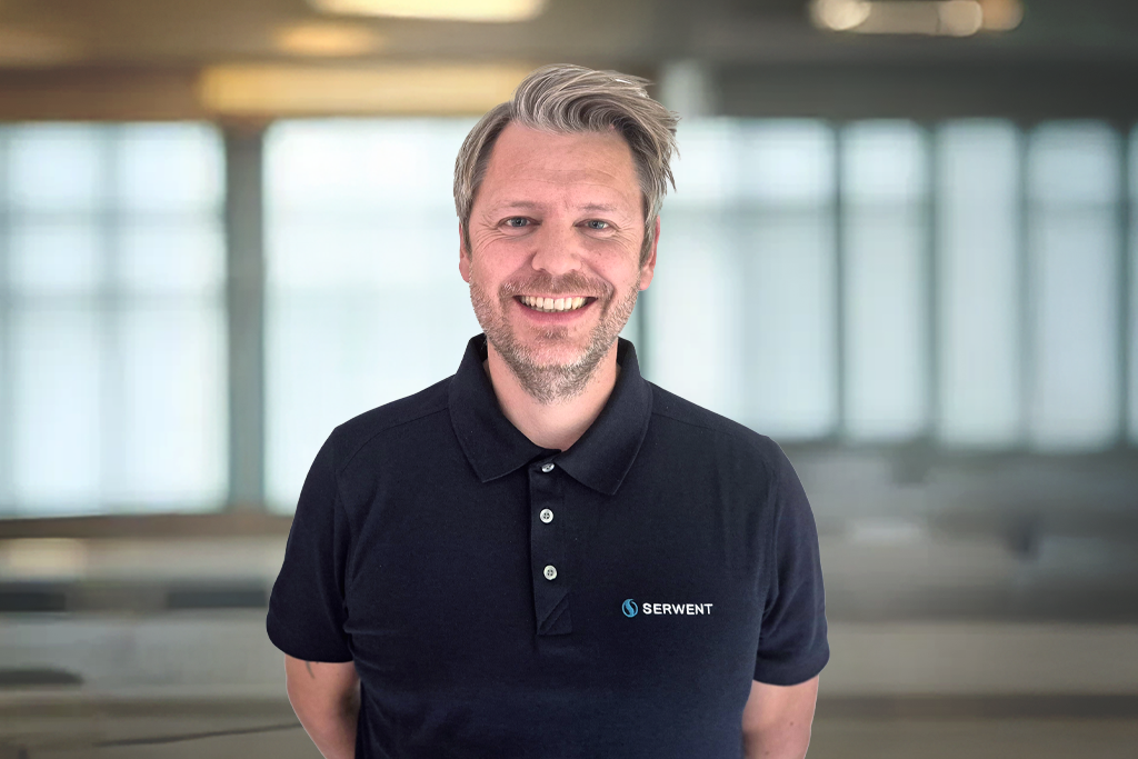 En mann med kort, lysebrunt hår og skjegg smiler og er iført en svart polo med SERWENT-logoen. Han står i en sterkt opplyst, uskarp innendørs kontorbakgrunn.