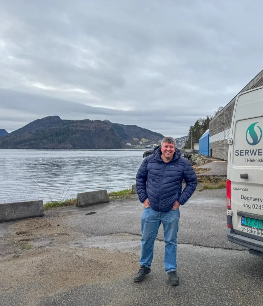 En mann i blå jakke og jeans står smilende nær en vannkant, ved siden av en hvit servicebil. Åser og bygninger er synlige over vannet under en overskyet himmel.