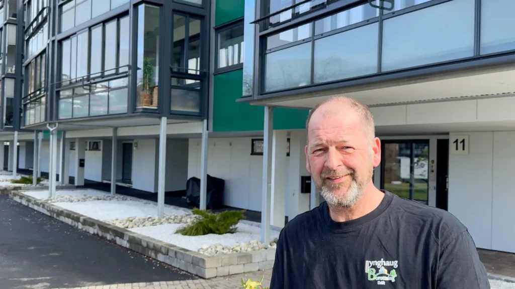 Serwent En middelaldrende mann med kort skjegg står utenfor moderne Lynghaug-leilighetsbygg med glassfasade, iført en svart t-skjorte og smiler til kameraet. Området rundt ham er pent anlagt med hvite steiner og små planter.