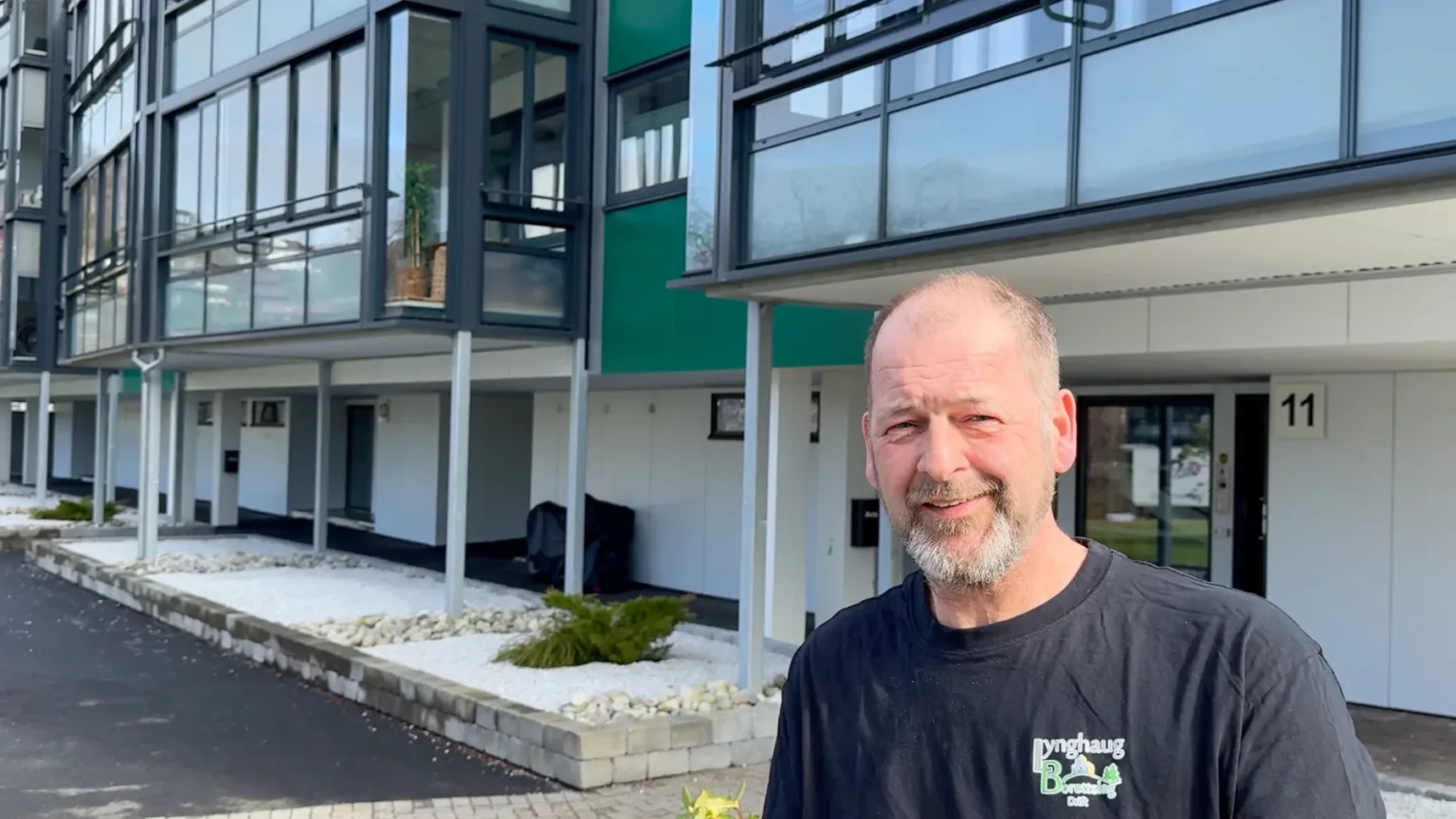 En middelaldrende mann med kort skjegg står utenfor moderne Lynghaug-leilighetsbygg med glassfasade, iført en svart t-skjorte og smiler til kameraet. Området rundt ham er pent anlagt med hvite steiner og små planter.