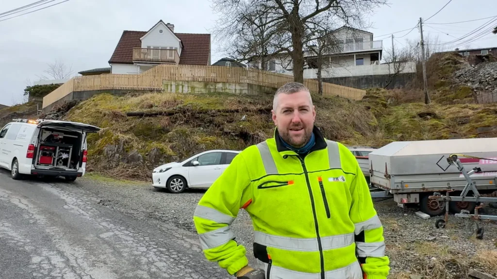En mann i en knallgul sikkerhetsvest står på en sprukket vei i nærheten av parkerte kjøretøy, med hus og et tregjerde i en gresskledd skråning i bakgrunnen på en overskyet dag.