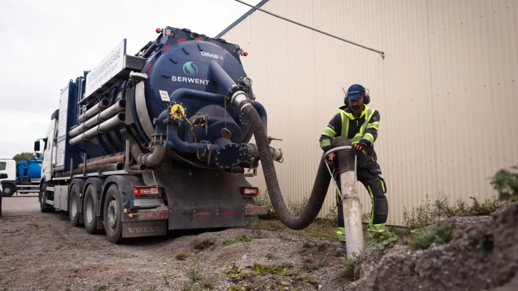 Supersuger En arbeider i synlig sikkerhetsutstyr bruker en stor støvsugerbil og holder en tykk slange for å suge opp materiale fra bakken ved siden av en beige industribygning.