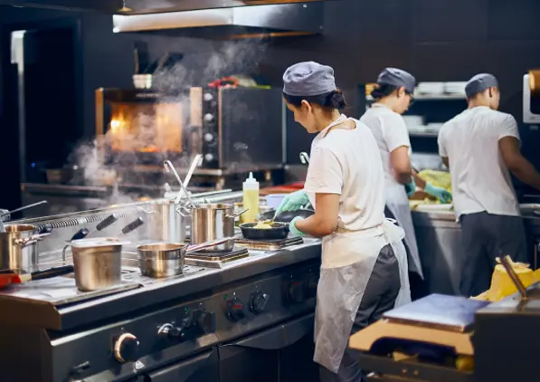 Tre kokker iført forklær og luer jobber i et travelt, moderne storkjøkken med gryter på komfyren, kjøkkenutstyr og damp som stiger opp mens de tilbereder mat.