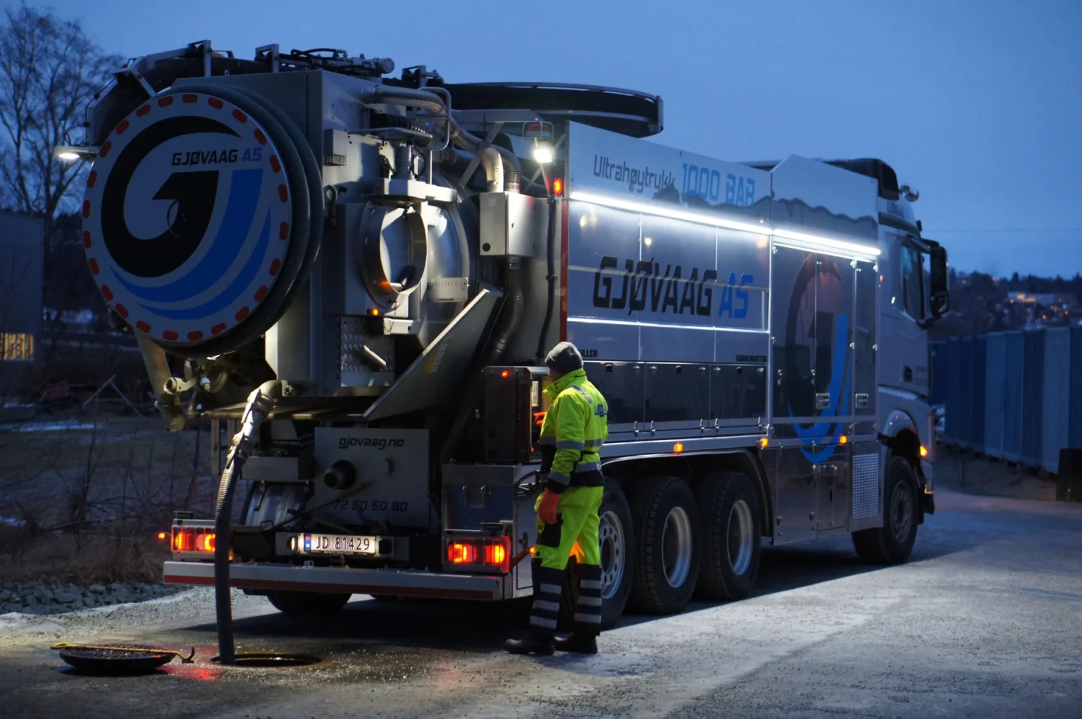 En arbeider iført varselutstyr betjener en stor støvsugerbil merket GJØVAAG AS i skumringen ved hjelp av en slange som er koblet til bakken i nærheten av et industriområde.