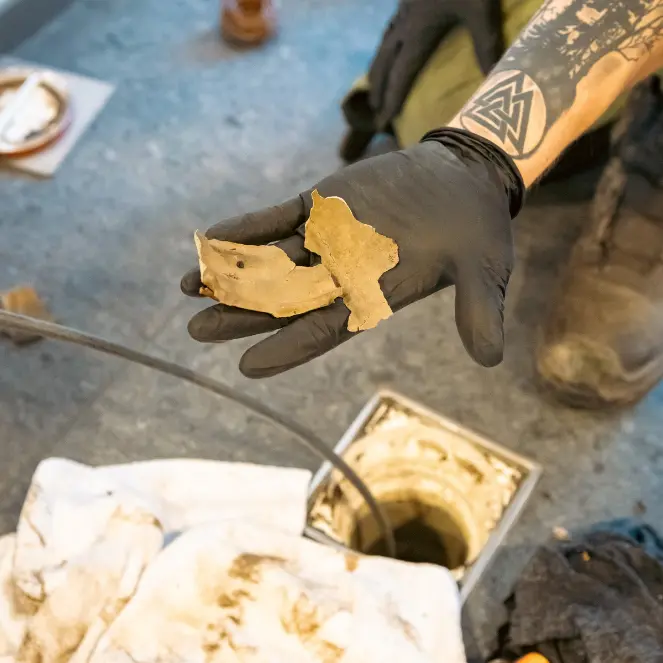 En person med svart hanske og tatovering holder opp en bit av noe som ser ut til å være et ødelagt rør eller kloakkfragment over et åpent avløp, med filler og rengjøringsverktøy i nærheten.