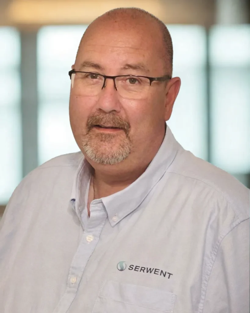 En middelaldrende mann med briller, fippskjegg og barbert hode bærer en lyseblå skjorte med Serwent-logoen, og står foran en uskarp kontorbakgrunn.