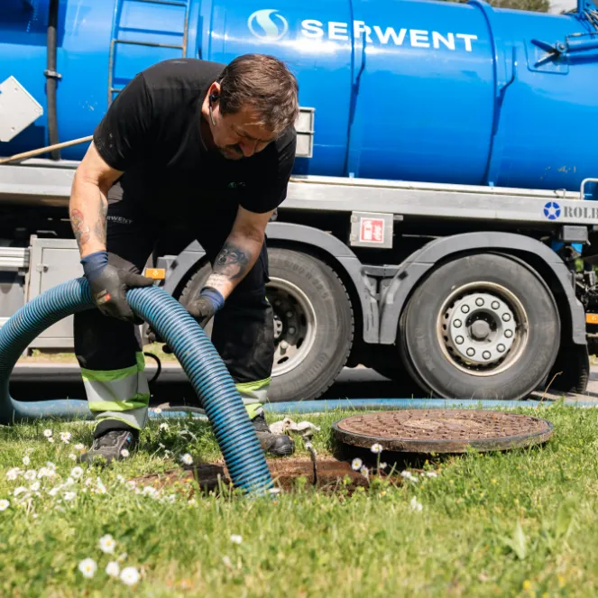En arbeider iført hansker og verneutstyr bruker en stor blå slange til septiktank- eller oljeutskillerkontroll, mens en blå SERWENT servicebil står parkert på gresset like ved.