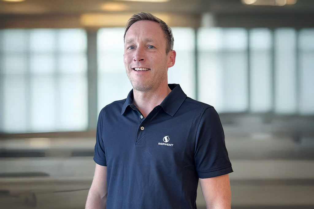 En smilende mann i marineblå poloskjorte med en liten logo står innendørs foran store, uskarpe vinduer.