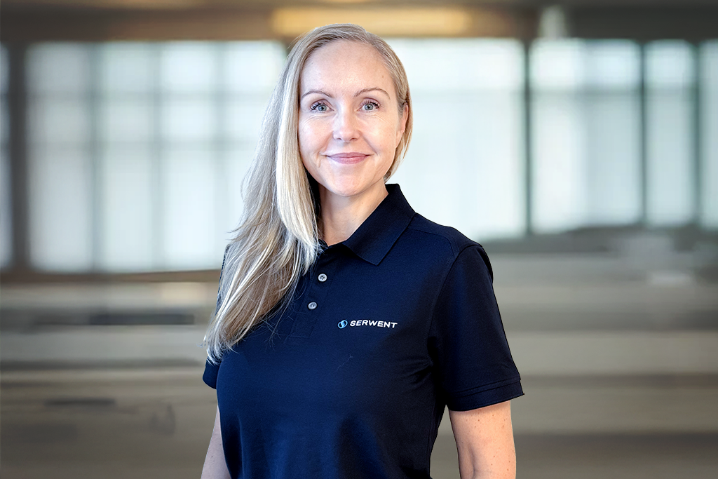 En kvinne med langt blondt hår og marineblå poloskjorte med SERVIENT-logo står innendørs i et lyst rom med store vinduer. Hun smiler og er vendt mot kameraet.