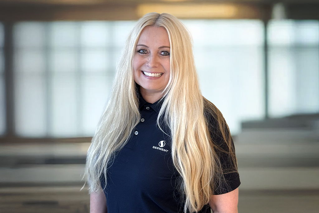 En smilende kvinne med langt, blondt hår og svart polo med Entrust-logo står i et sterkt opplyst kontormiljø.