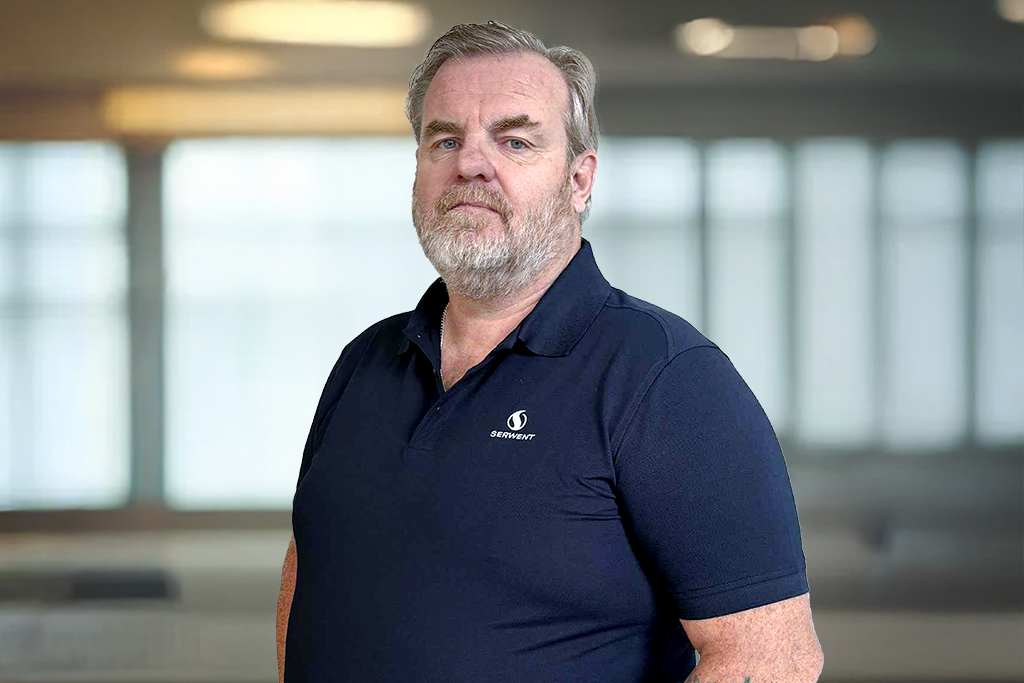 En middelaldrende mann med grått hår og skjegg står innendørs, iført en marineblå poloskjorte med logo på brystet. Bakgrunnen er uskarp, og viser store vinduer og sterk innendørsbelysning.