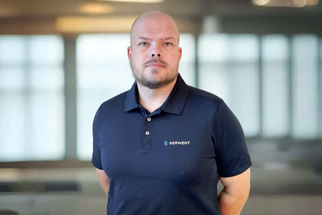 En mann med barbert hode og skjegg, iført marineblå skjorte med Serwent-logo, står innendørs foran store vinduer med uskarpt naturlig lys.