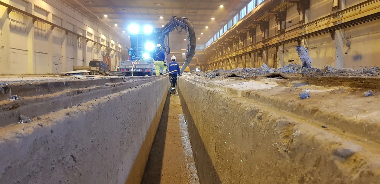 Arbeidere betjener anleggsmaskiner inne i en stor industribygning, sett fra mellom to betongspor. Sterkt lys lyser opp scenen og fremhever støv og rusk på betongoverflatene.