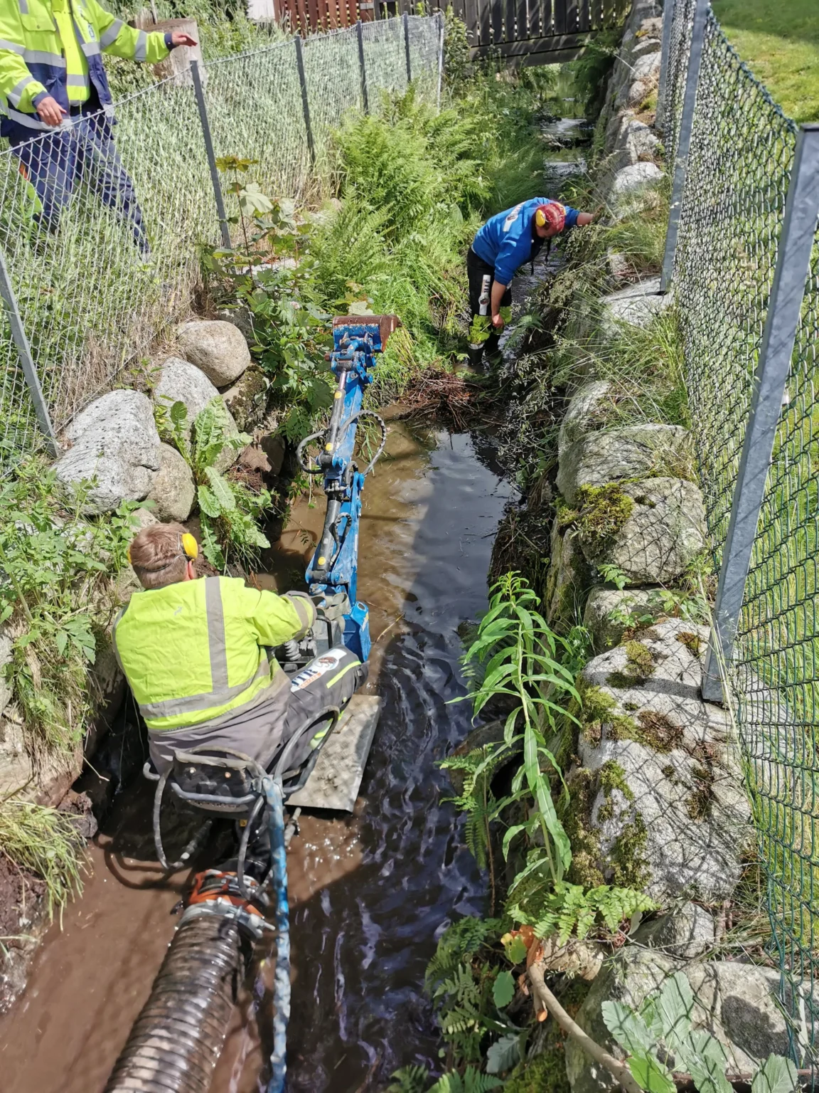 To personer arbeider i et smalt, steinete elveleie mellom nettinggjerder. Den ene betjener en liten blå maskin i vannet, mens den andre samler søppel for hånd. Langs bredden vokser det bregner og grønne planter.