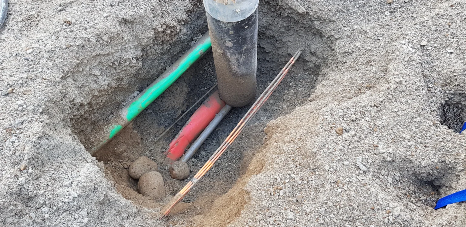 I et grunt hull i sandjorda ligger det flere underjordiske ledninger, blant annet grønne, røde og kobberledninger, ved siden av en vertikal, sylindrisk stolpe som er delvis nedgravd i bakken.