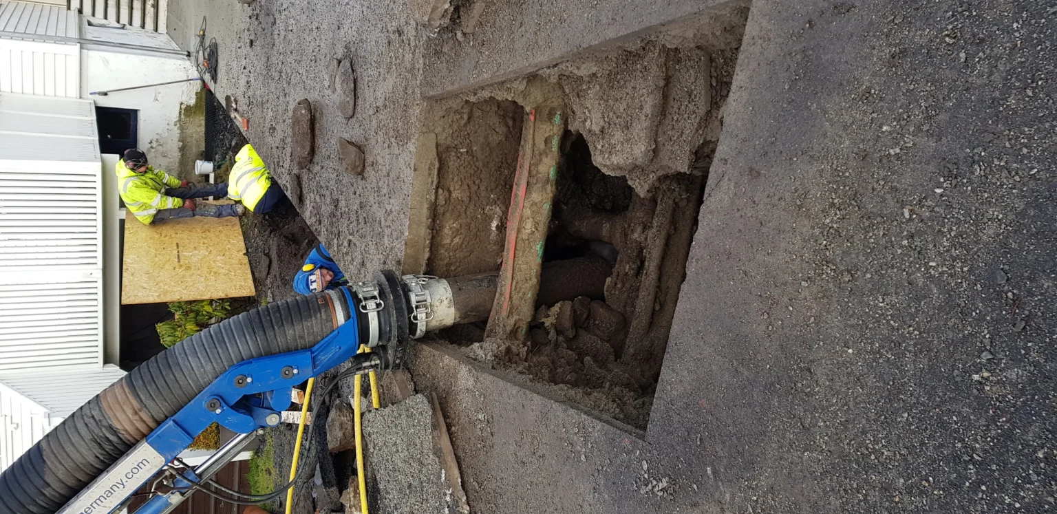 To arbeidere i gult verneutstyr befinner seg på en byggeplass med et stort rektangulært hull i bakken og en blå vakuumgravemaskin som fjerner jord. Rundt hullet ligger asfaltbiter og jord.