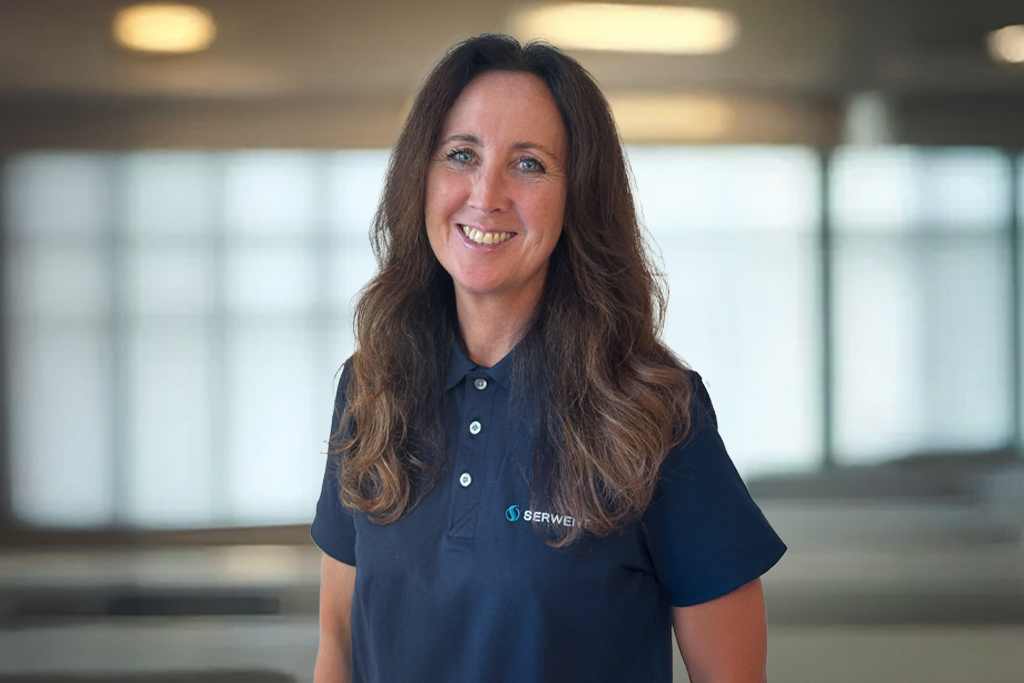 En kvinne med langt, brunt hår smiler i en marineblå poloskjorte med SERWEL-logo, mens hun står innendørs i et lyst, moderne kontorlokale.