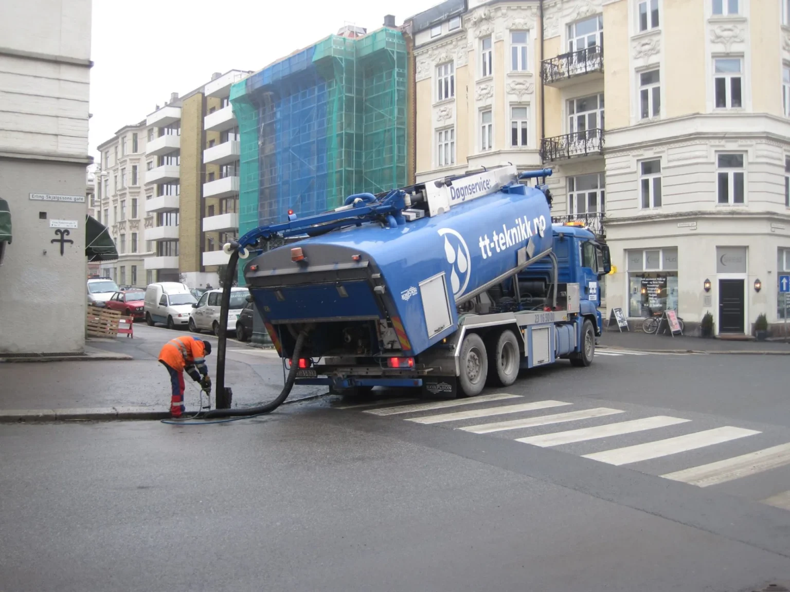 En arbeider i oransje vernetrøye utfører avloppsrensing med en slange koblet til en blå forsyningsbil som står parkert i en bygate nær et fotgjengerfelt, med boligblokker og stillaser i bakgrunnen.