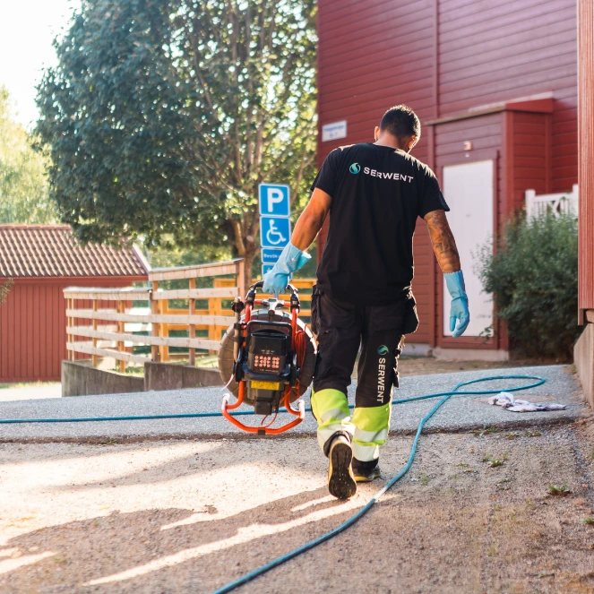 En arbeider iført hansker og Serwent-skjorte går utendørs i nærheten av en rød bygning og bærer utstyr med kabler på en grussti. Et blått parkeringsskilt for funksjonshemmede er synlig i bakgrunnen.