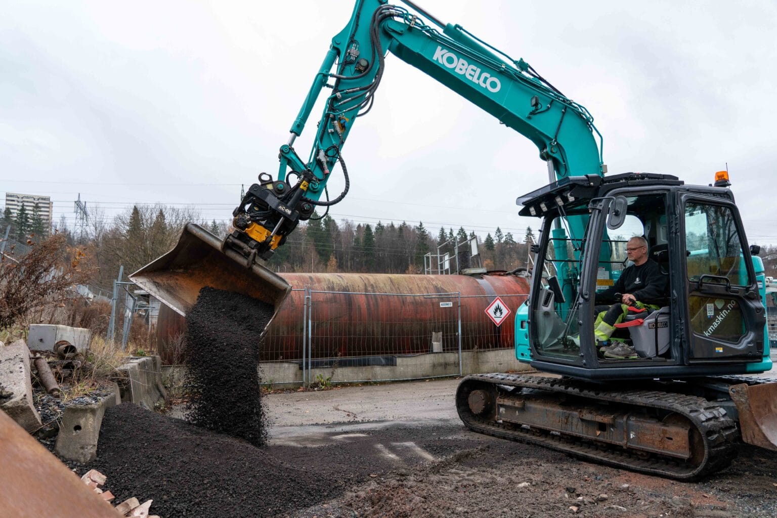 En person kjører en blå Kobelco-gravemaskin og bruker skuffen til å helle grus eller asfalt på en byggeplass omgitt av gjerder, rustne tanker og trær i bakgrunnen.