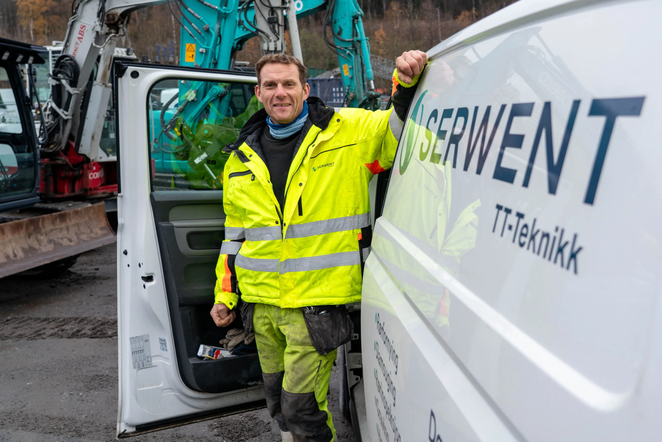 En mann i gul refleksjakke står smilende ved siden av en åpen varebildør med "SERVE NT IT-Teknikk" skrevet på. I bakgrunnen skimtes anleggskjøretøy og trær.