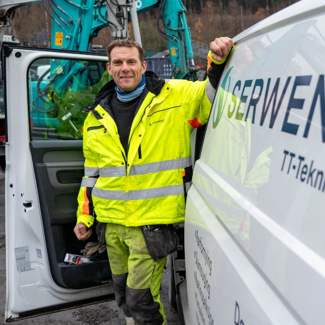 En anleggsarbeider i knallgul jakke står og smiler ved siden av en hvit servicebil med åpen dør. I bakgrunnen ser vi blå anleggsmaskiner og et kupert landskap.