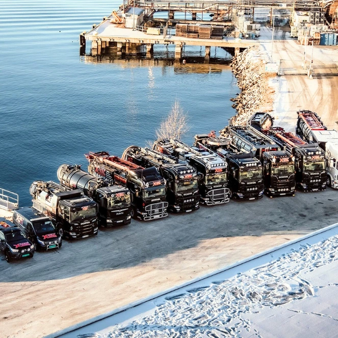 En rekke store, svarte lastebiler og tunge kjøretøy står parkert side om side på en snødekt elvebredd, i nærheten av en brygge som strekker seg ut i stille, blått vann under en klar himmel.