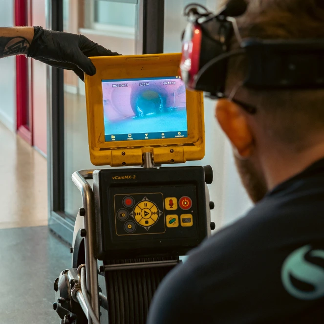En person iført hansker og verneutstyr betjener et rørinspeksjonskamera og ser på innsiden av et rør på en skjerm som er festet til utstyret. Skjermen viser et blåtonet bilde av rørets innside.