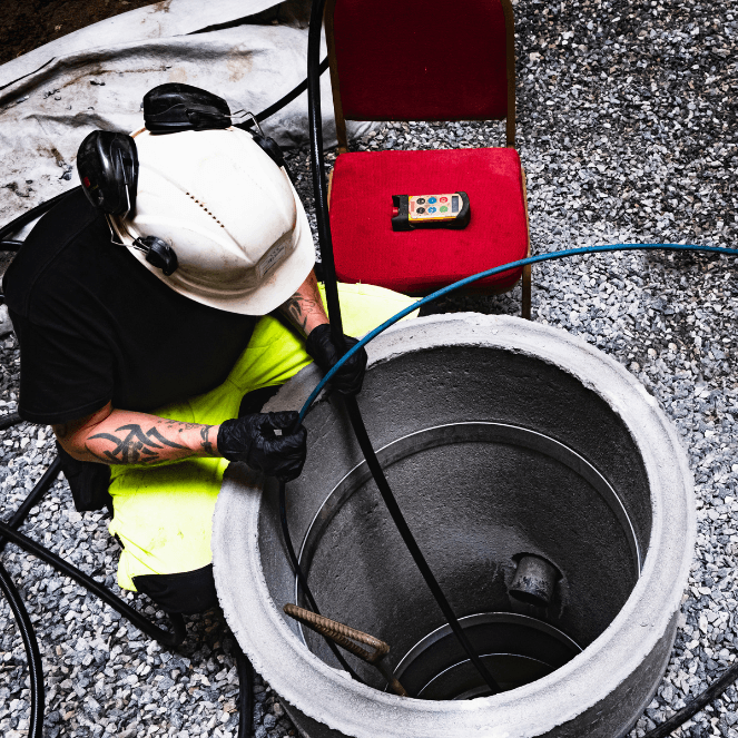 En anleggsarbeider i verneutstyr håndterer kabler i nærheten av en åpen betongkum, med en rød stol og et høytrykksspylingapparat på bakken like ved. Grus dekker området rundt.