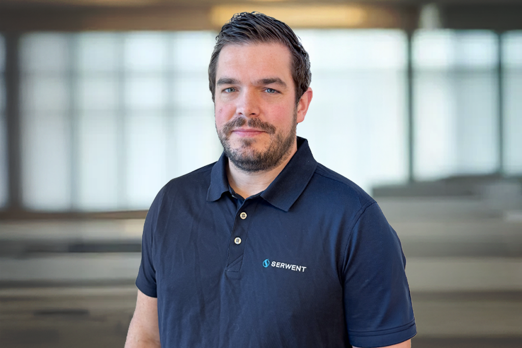 En mann med kort, mørkt hår og skjegg, iført marineblå SERWENT-genser, står på et kontor med store vinduer og uskarp bakgrunn.