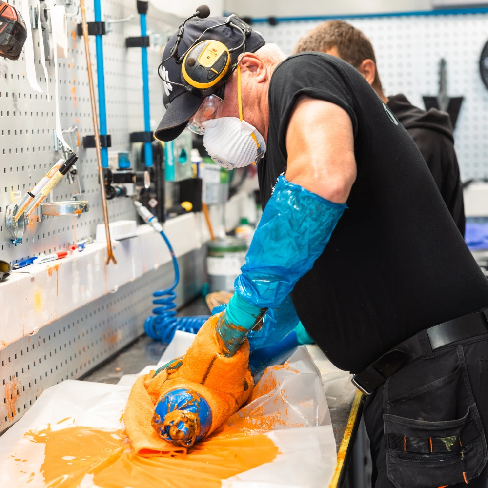 En person iført verneutstyr og blå hansker vikler et oransje tøystykke rundt en sylindrisk gjenstand, muligens relatert til garanti rørfornying, ved en verkstedbenk med diverse verktøy på veggen.