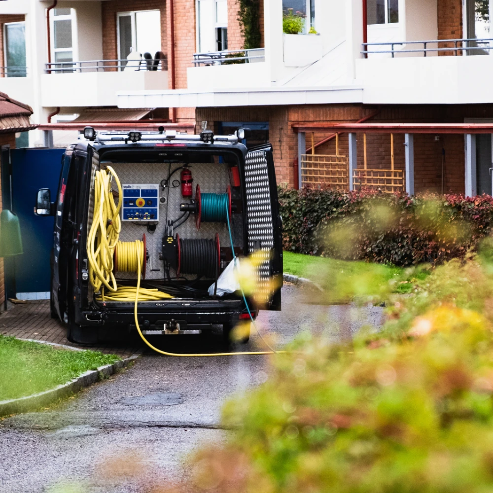 En varebil med åpne bakdører og opprullede slanger står parkert i en oppkjørsel nær en boligblokk, noe som tyder på at det pågår vedlikeholds- eller reparasjonsarbeid. Busker og grøntområder omkranser området.