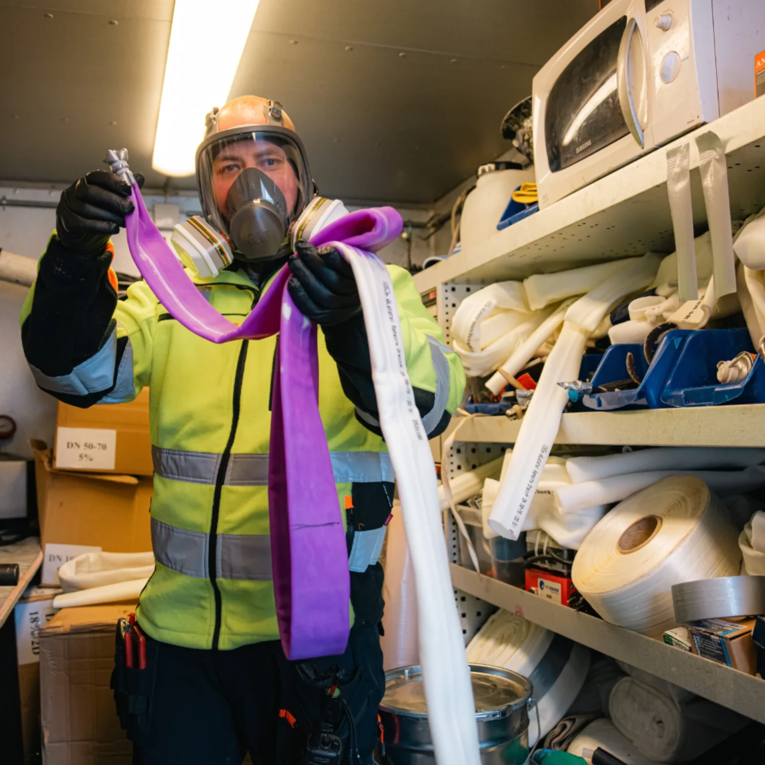 En person i verneutstyr og åndedrettsvern holder to slanger, en lilla og en hvit, inne i et lagerrom fylt med slanger, verktøy og en mikrobølgeovn på en hylle.