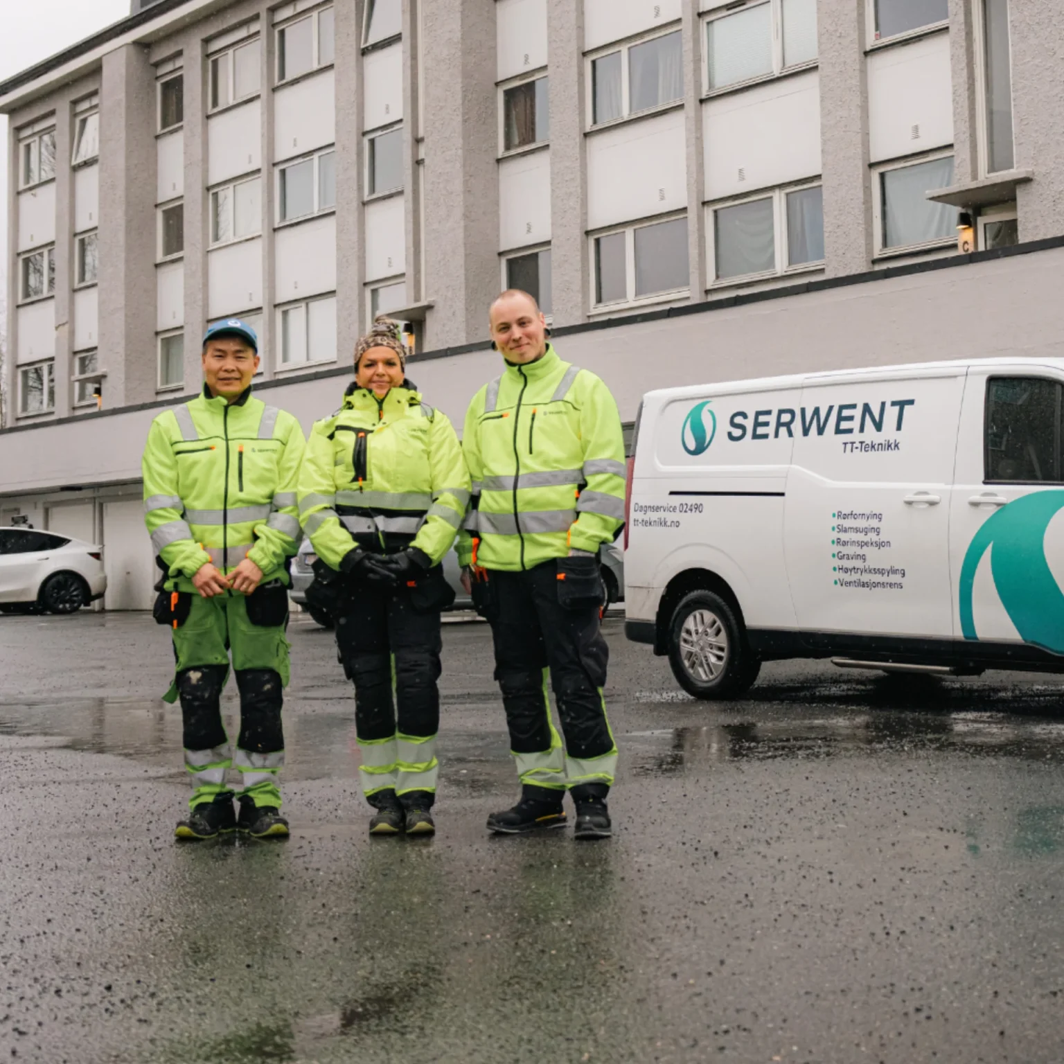 Tre personer i godt synlige arbeidsklær står sammen og smiler foran en bygning og en hvit varebil merket SERWENT på en våt gate.