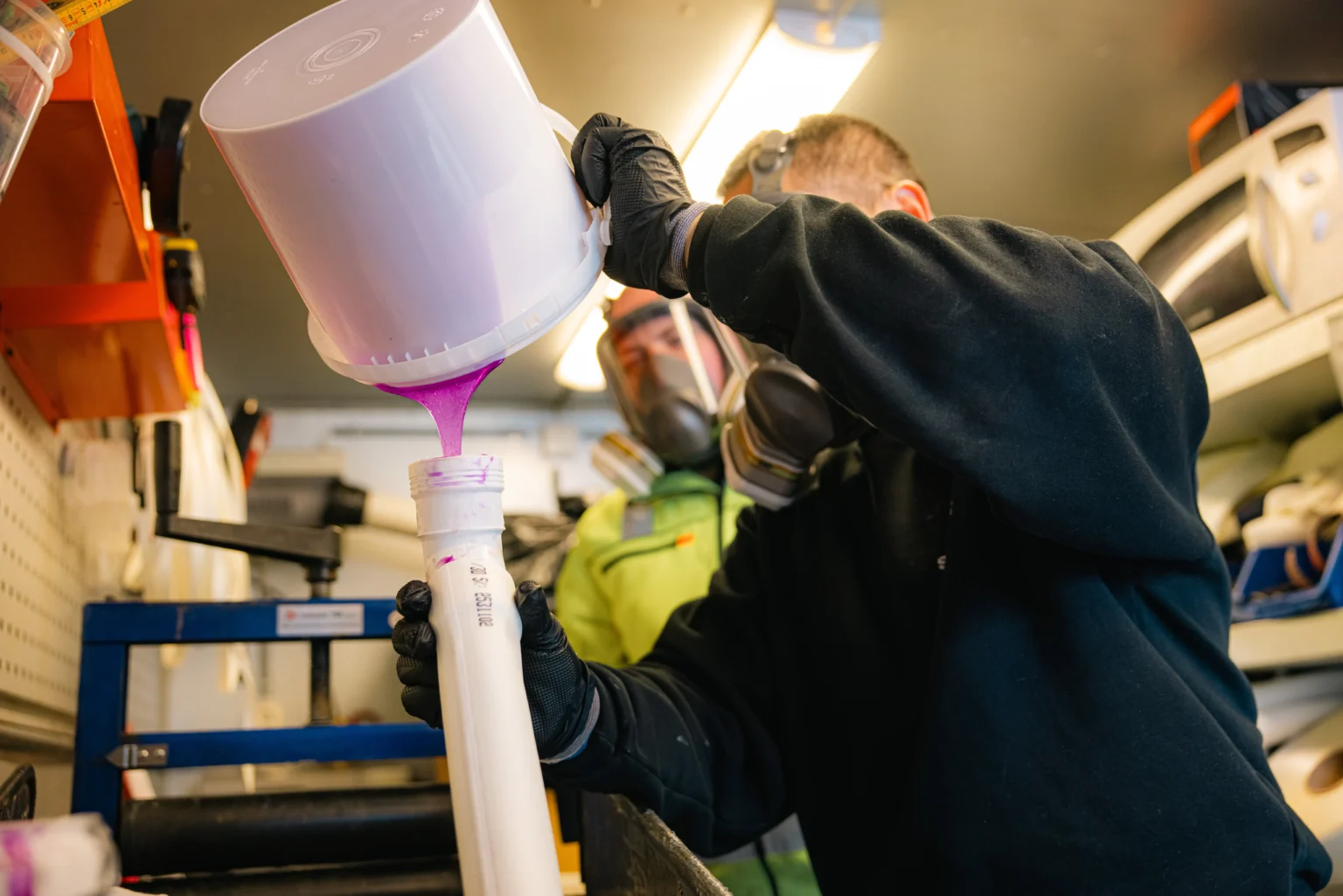 En person som har på seg hansker og åndedrettsmaske, heller en knallrosa væske fra en bøtte ned i et høyt, smalt rør i et verksted, og demonstrerer dermed et viktig trinn i rørfornyingsprosessen.