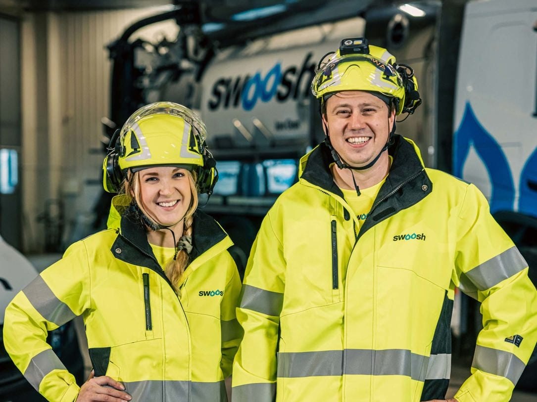 To personer i gule refleksjakker og hjelmer smiler mens de står innendørs. Bak dem kan man delvis se et stort kjøretøy med påskriften «SWOOSH».