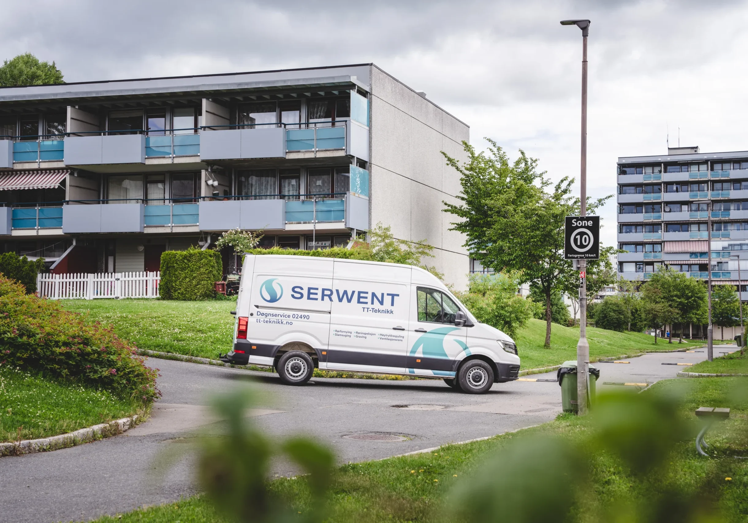 En hvit varebil med logoen «Serwent» står parkert i en boliggate ved siden av en moderne bygård. Frodig grønt gress og trær rammer inn scenen, mens et fartsgrenseskilt på 10 kompletterer den overskyede himmelen, og gjenspeiler Serwents forpliktelse til å forbedre urbant inneklima.