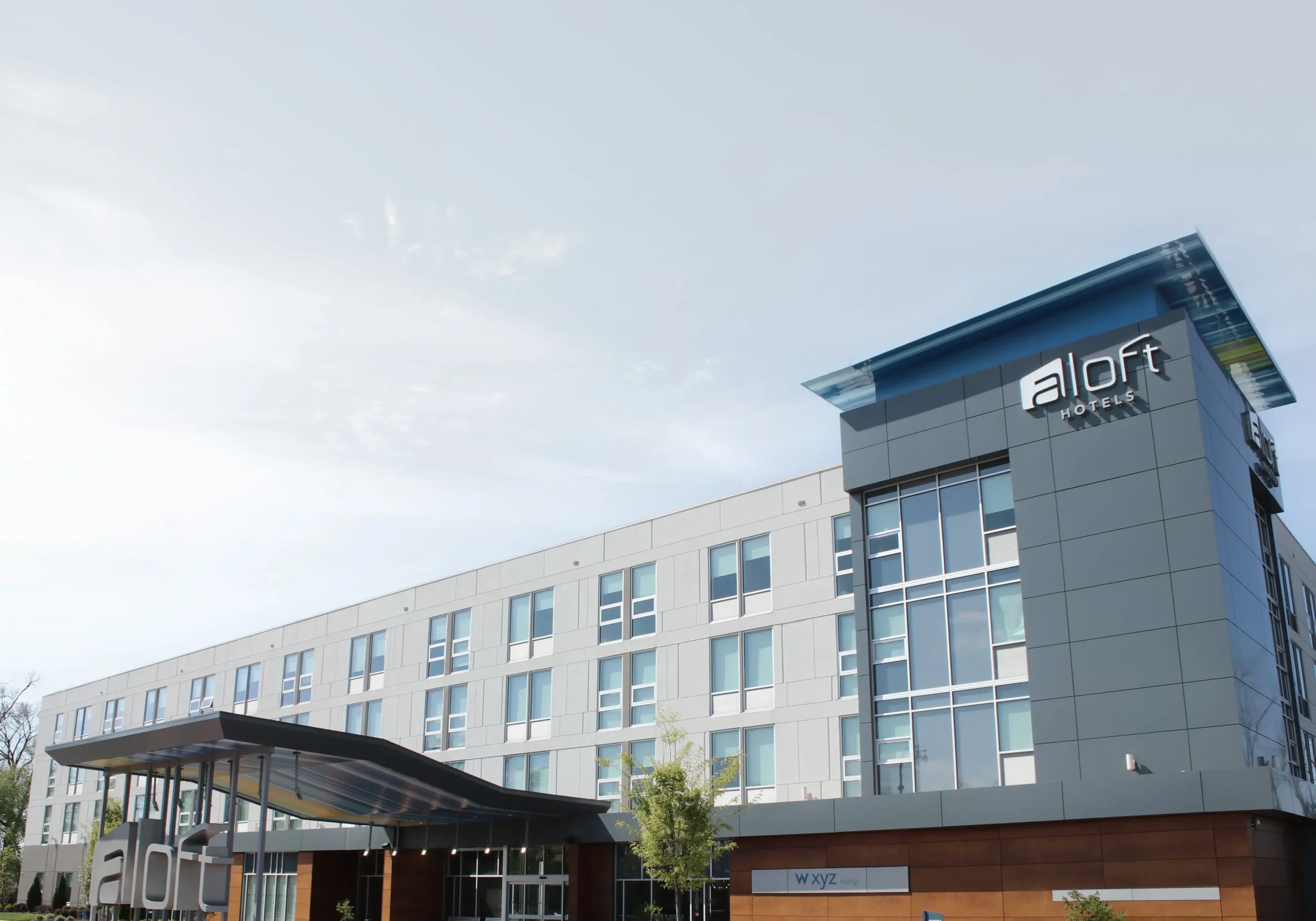 En moderne Aloft Hotel-bygning i flere etasjer med store vinduer, en elegant inngangsbaldakin og anlagte omgivelser under en delvis overskyet himmel.