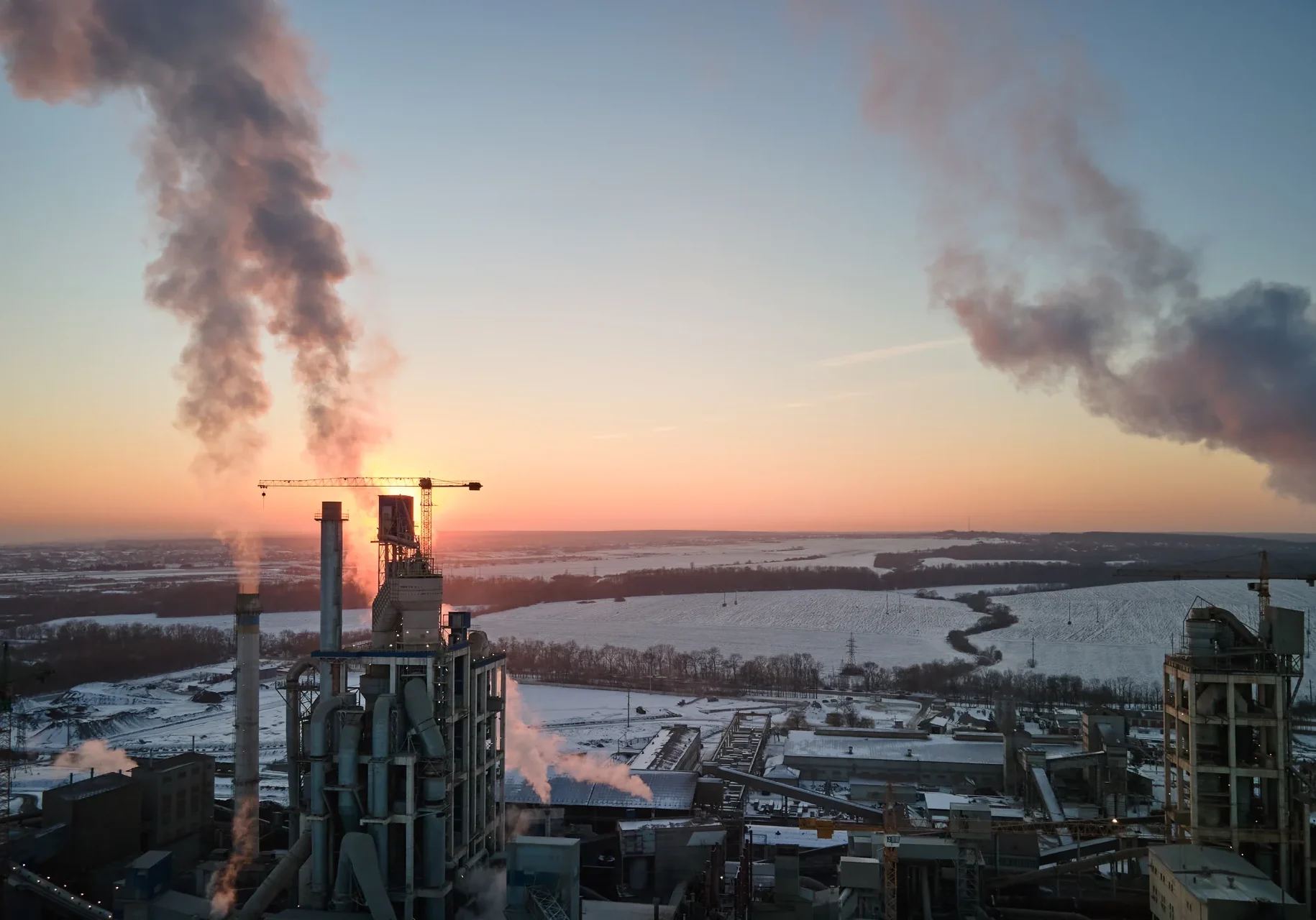 Industrianlegg med høye skorsteiner som slipper ut røyk, plassert mot et vinterlandskap med snødekte jorder og fjerne trær ved soloppgang eller solnedgang. Himmelen er klar, og sollyset filtrerer gjennom røyken.