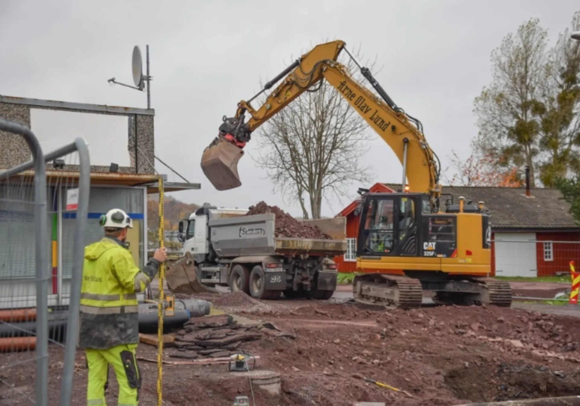 En byggeplass der entreprenører jobber effektivt: En arbeider i gul sikkerhetsdrakt måler bakken mens en gravemaskin laster jord opp i en dumper. Bygninger og trær skimtes i bakgrunnen.